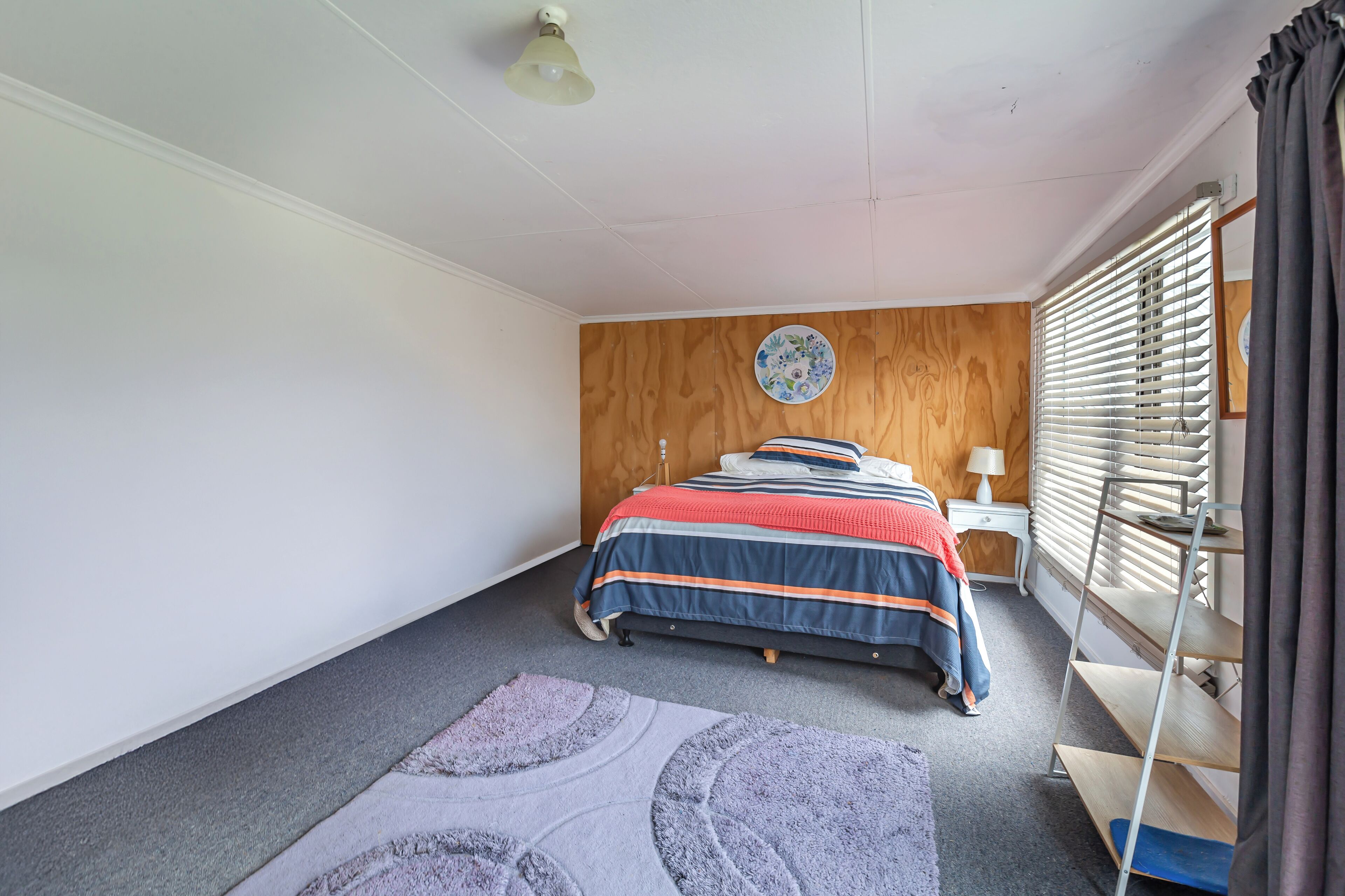 Une chambre à coucher comprenant un lit, une table de chevet, une lampe, une tête de lit en bois, une assiette décorative accrochée au mur et une fenêtre avec des stores.