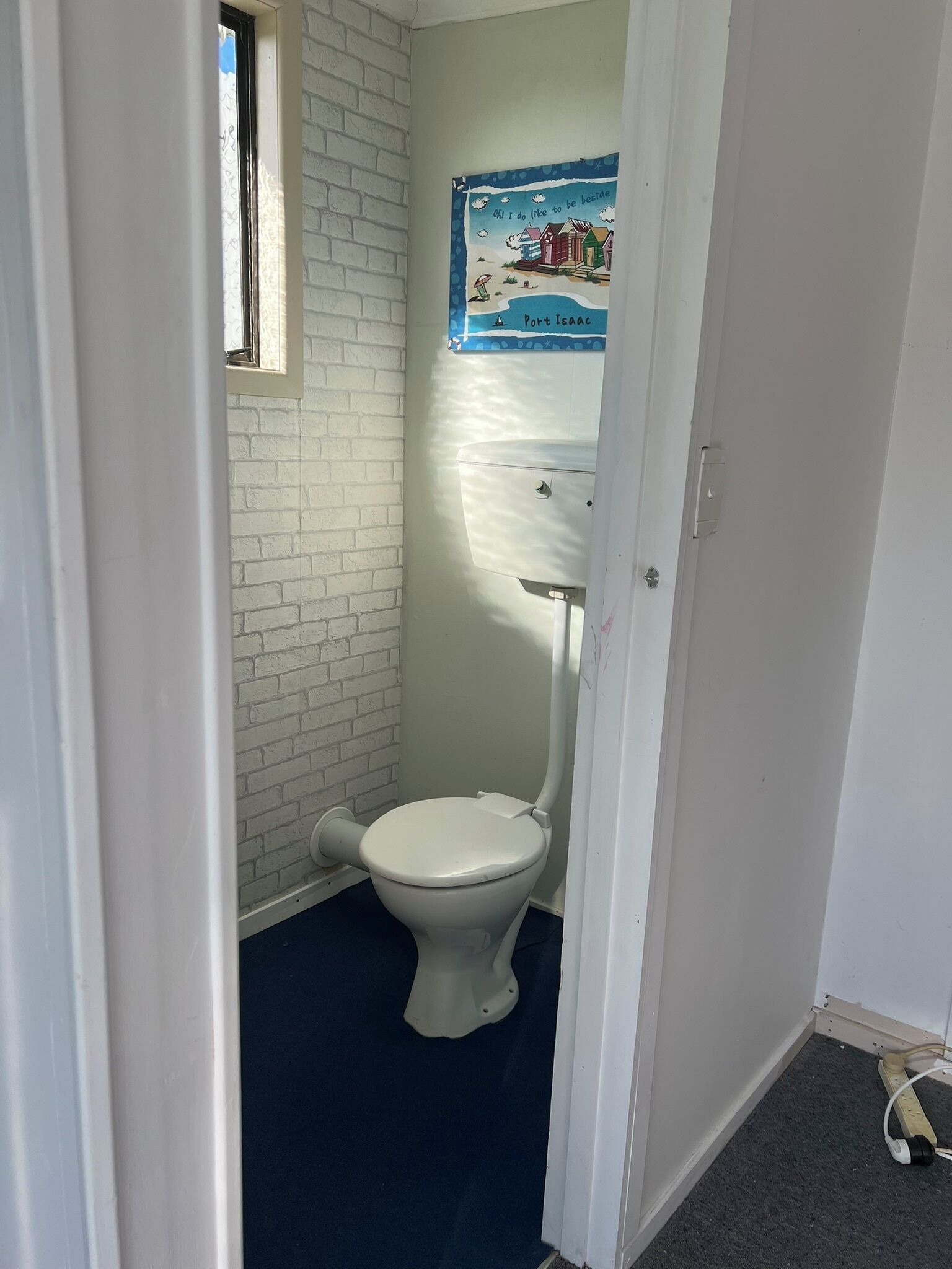 A small, narrow bathroom with a white toilet, a window, and a poster on the wall.