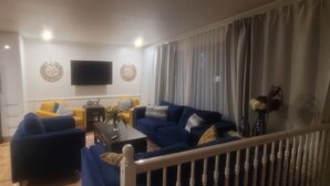 Una sala de estar con un sofá azul, un sofá amarillo, un televisor de pantalla plana montado en la pared y un ventanal con cortinas.
