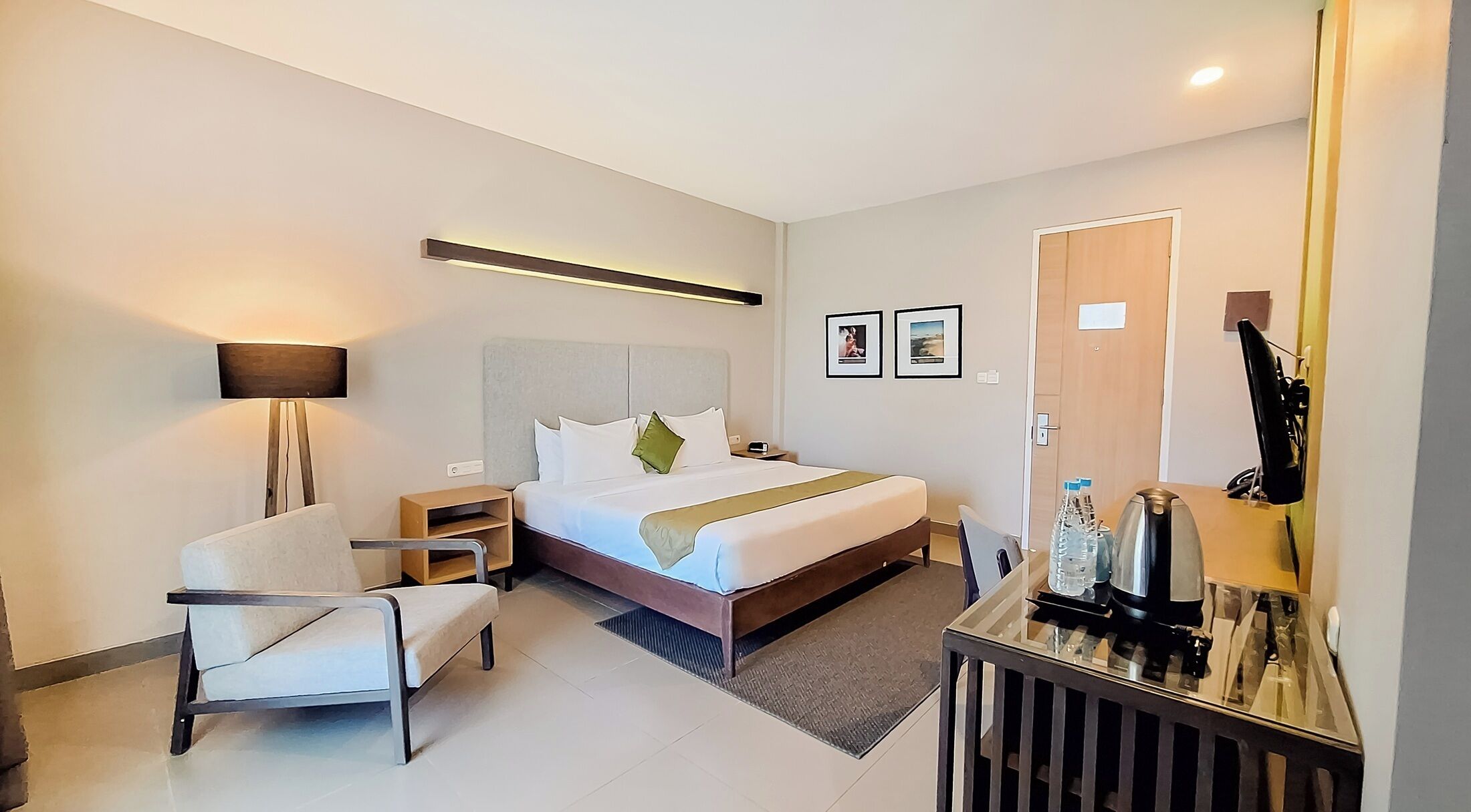 Une chambre d’hôtel moderne équipée d’un lit, d’une table de chevet, d’une chaise, d’une lampe et d’un bureau avec un miroir et des bouteilles d’eau.