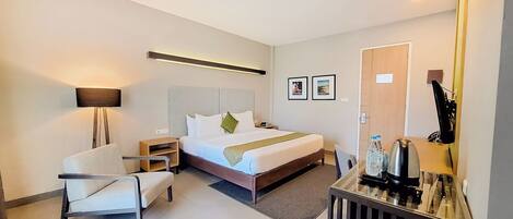 Una habitación de hotel moderna con cama, mesita de noche, silla, lámpara y escritorio con espejo y botellas de agua.