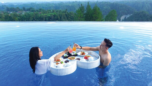 Two people toasting with drinks in a large, clear pool with a scenic view of trees and hills in the background. - The Balcone Hotel & Resort (Bukittinggi)