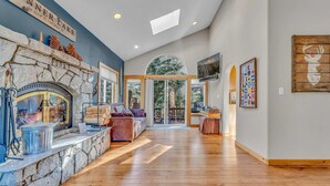 A spacious living area with a stone fireplace, wooden flooring, and a large window overlooking a deck. - Spacious Pet-Friendly Truckee Retreat with Hot Tub (Truckee)