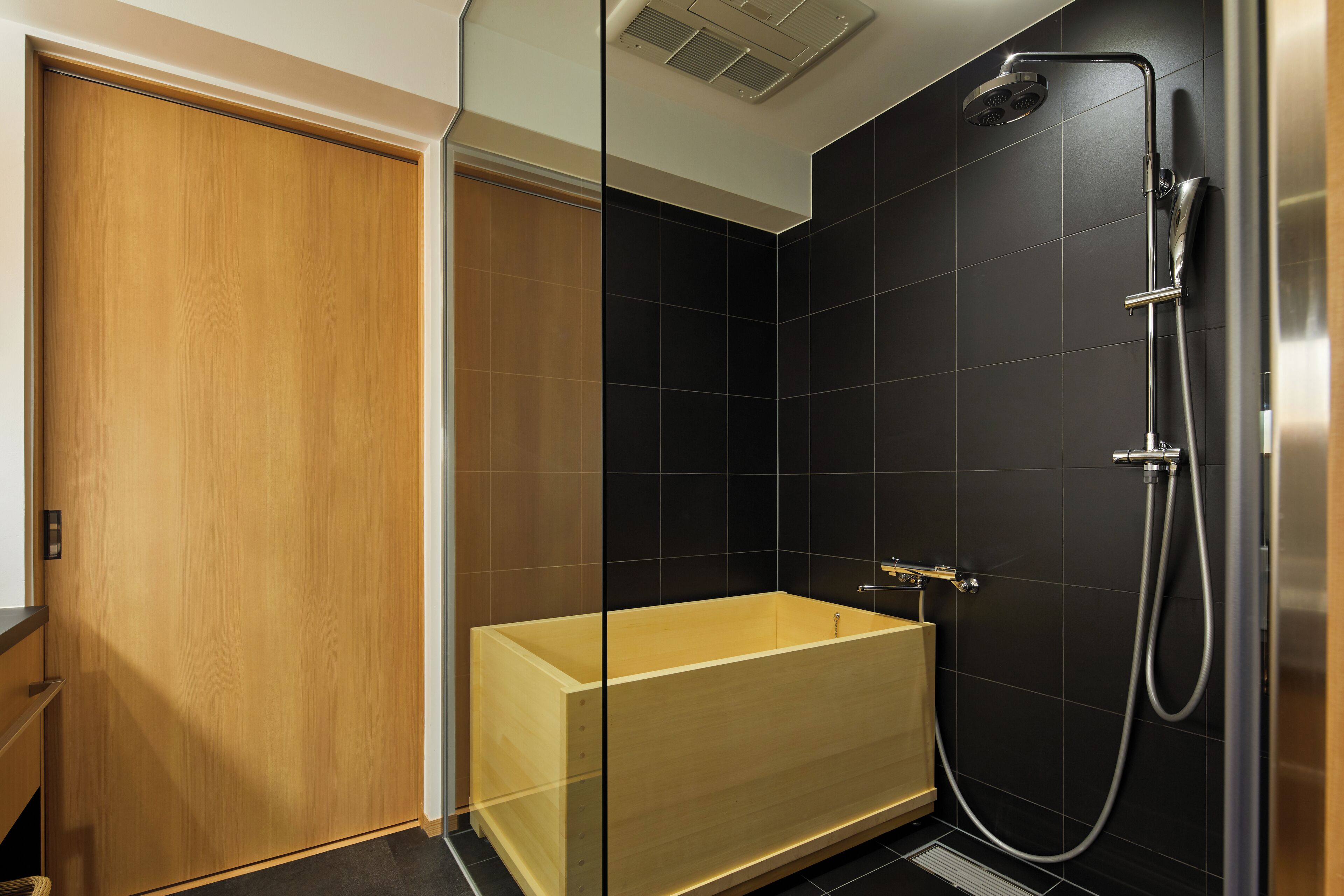 A modern bathroom with a wooden bathtub, a shower with a handheld hose, and black tiled walls.