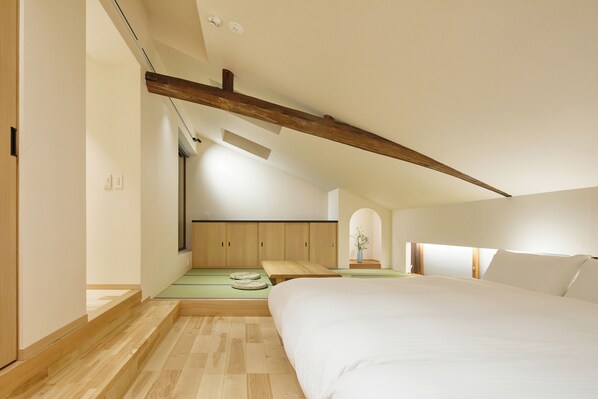 Quarto com cama grande, vigas de madeira, um pequeno espaço de estar e vista para um jardim.