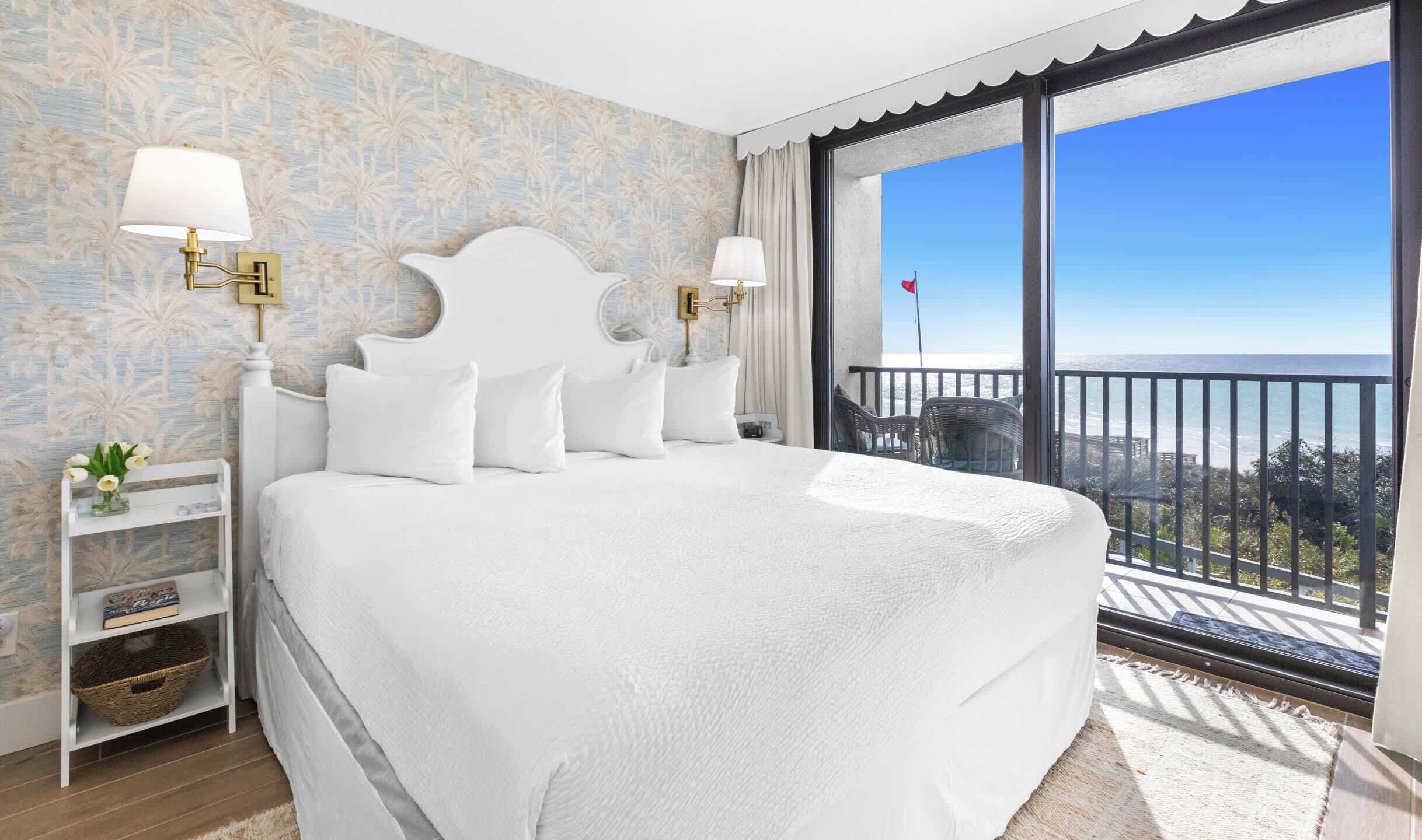 A large bed with white bedding, two bedside lamps, a nightstand with a vase and books, and a view of the ocean through a balcony with a railing.