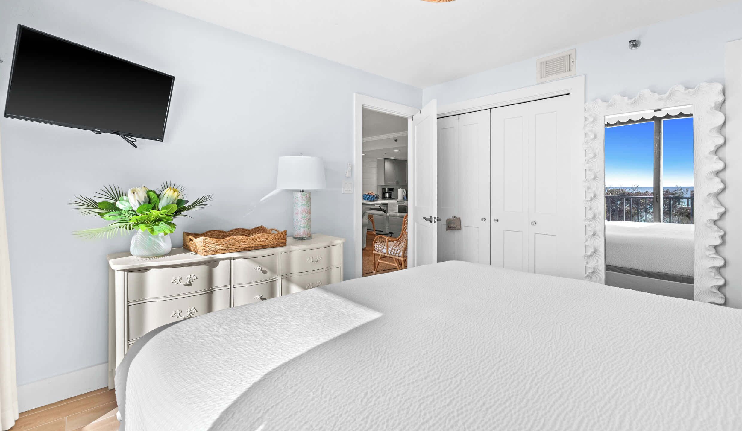 Een slaapkamer met een groot bed, een aan de muur bevestigde televisie, een dressoir met lades, een rieten mand en een bloempot.