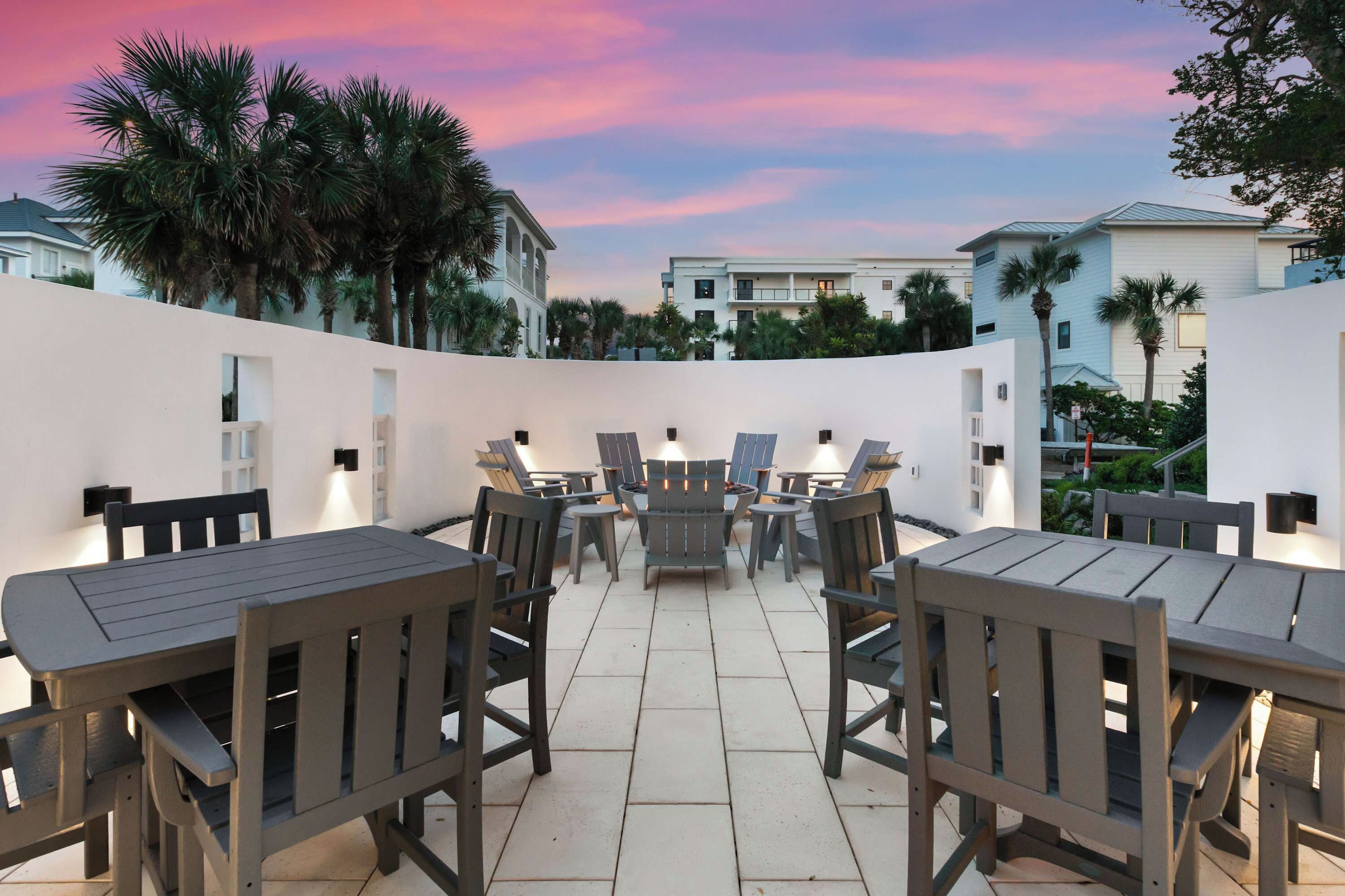 An outdoor patio with wooden tables and chairs, surrounded by a white wall and palm trees.