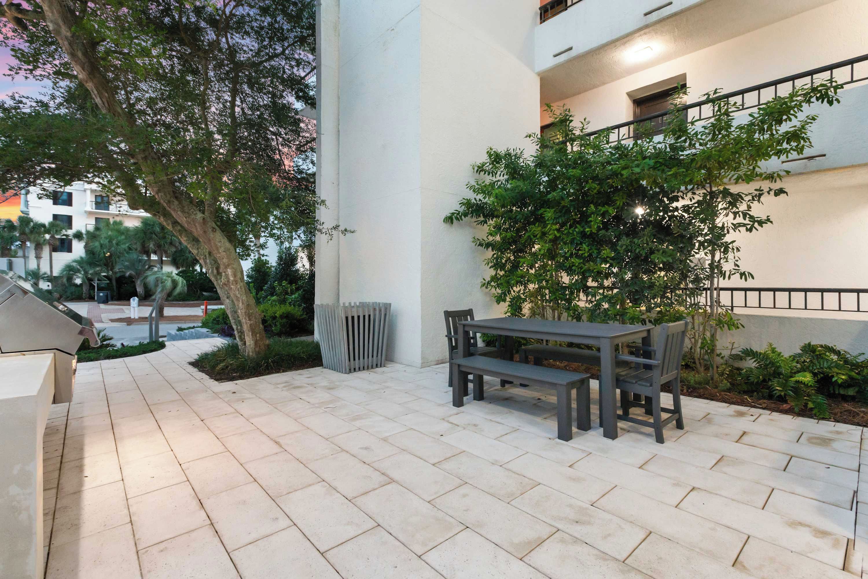 An outdoor patio area with a table and benches, surrounded by greenery and a white building.