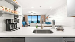 A modern kitchen with a built-in dishwasher, a coffee maker, and a large window overlooking the sea.