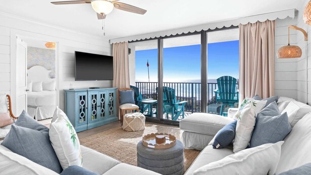 A spacious living room with a large TV, comfortable seating, a balcony with a view, and a ceiling fan.