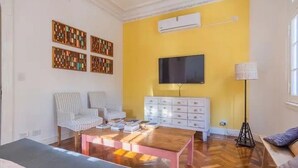 Una sala de estar con pared amarilla, un televisor de pantalla plana, un armario blanco, una silla a rayas, una mesa de centro de madera y una lámpara de pie.