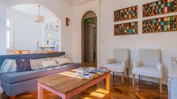 Una habitación con un sofá, dos sillas, una mesa de madera con libros y una pared decorada con obras de arte coloridas.
