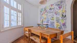 Un comedor con una mesa de madera y bancos, un ventanal con contraventanas blancas y una pared decorada con varios adhesivos y dibujos.