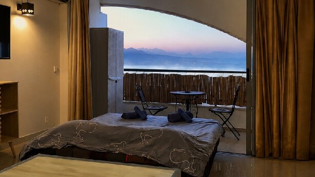 Une chambre d’hôtel avec un lit, une petite table, une chaise et un balcon offrant une vue sur la mer et les montagnes.