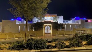 Un bâtiment avec un grand arbre devant, une entrée fermée par un portail et une enseigne lumineuse.