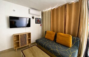Uma sala de estar com televisão de tela plana, um rack de madeira, sofá com almofadas e uma janela com cortinas.