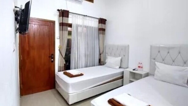 A hotel room with two beds, a wooden door, a window with curtains, a bedside table with a lamp, and a wall-mounted air conditioning unit. - Sandi Guest House Syariah (Banyuwangi)