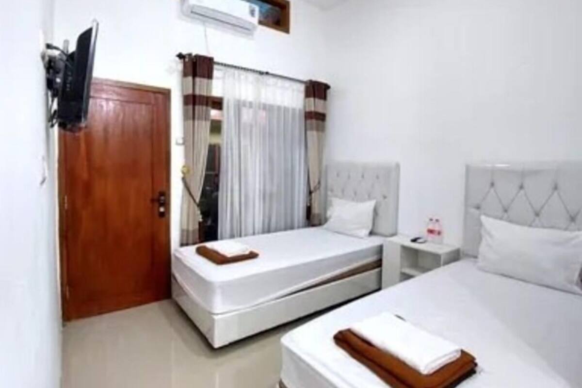 A hotel room with two beds, a wooden door, a window with curtains, a bedside table with a lamp, and a wall-mounted air conditioning unit.