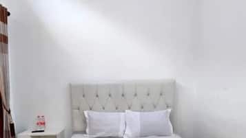 Una habitación de hotel con una cama bien tendida, una mesita de noche pequeña con una botella y una pared blanca lisa.