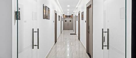 A long, narrow hallway with white walls and doors on both sides, leading to a central area with a door at the end.