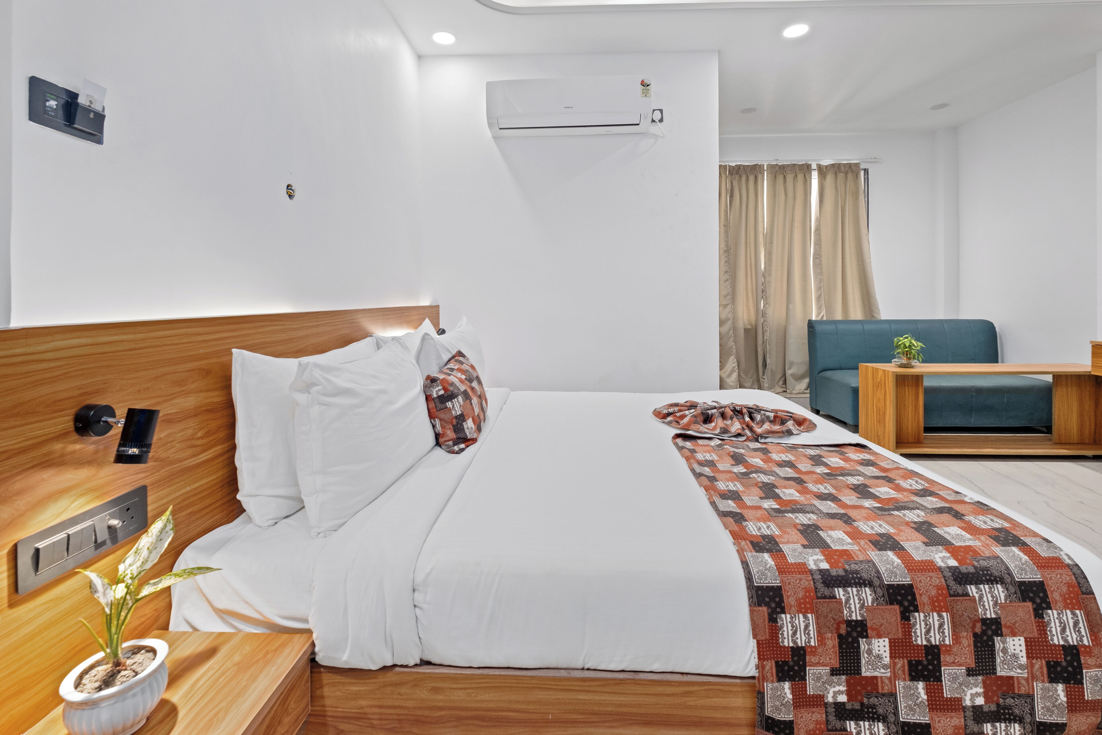 Habitación de hotel con cama, mesita de noche con una planta, escritorio con silla, sofá azul y unidad de aire acondicionado.