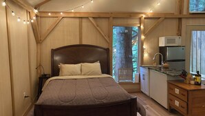 A cozy bedroom with a wooden bed, a nightstand, and a small desk with a microwave. A window offers a view of a tree outside. - Unique Cortes Treehouse! - Peaceful haven in the trees (Strathcona B)