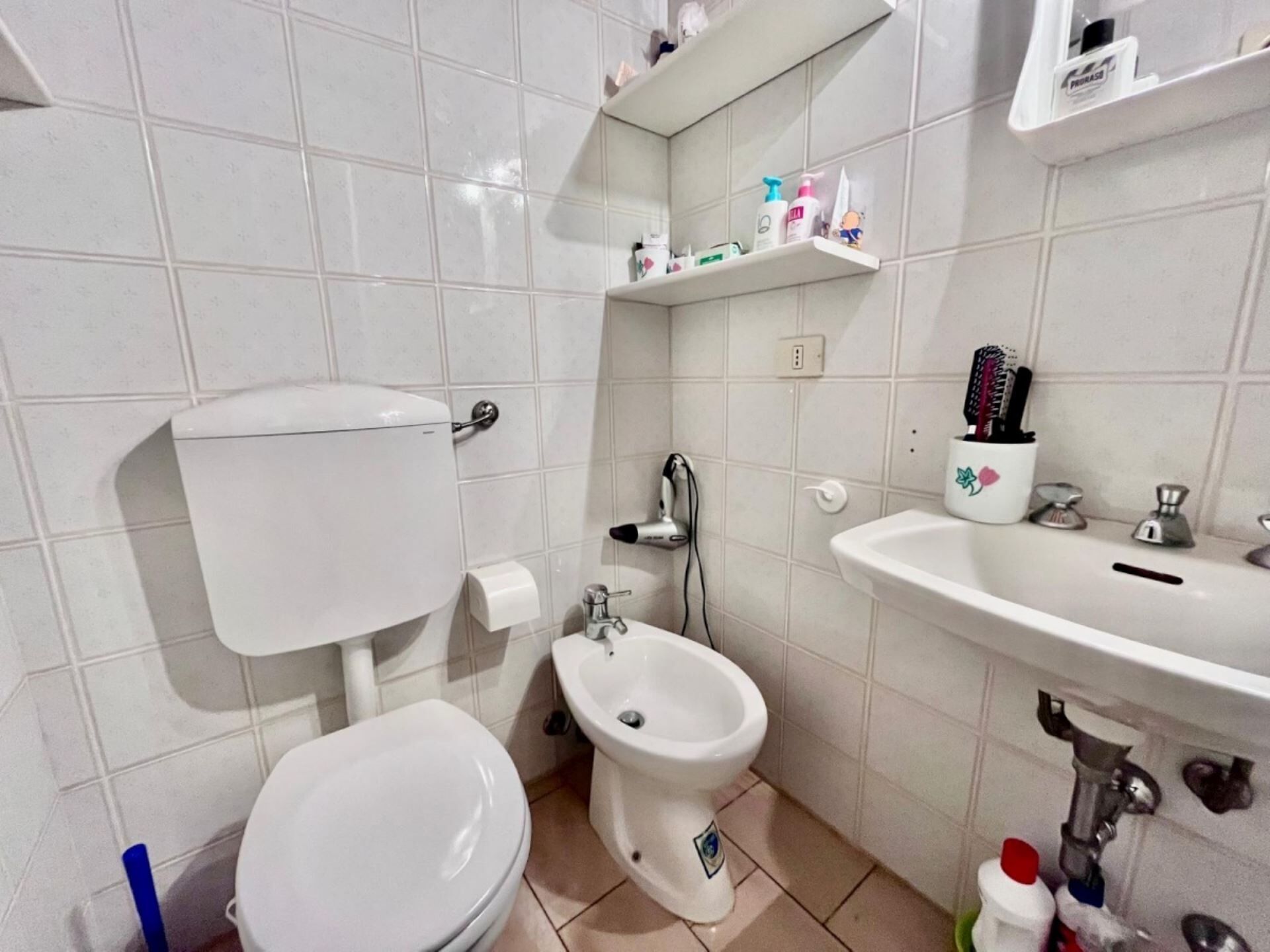 A bathroom with a toilet, bidet, sink, and shelves with toiletries.