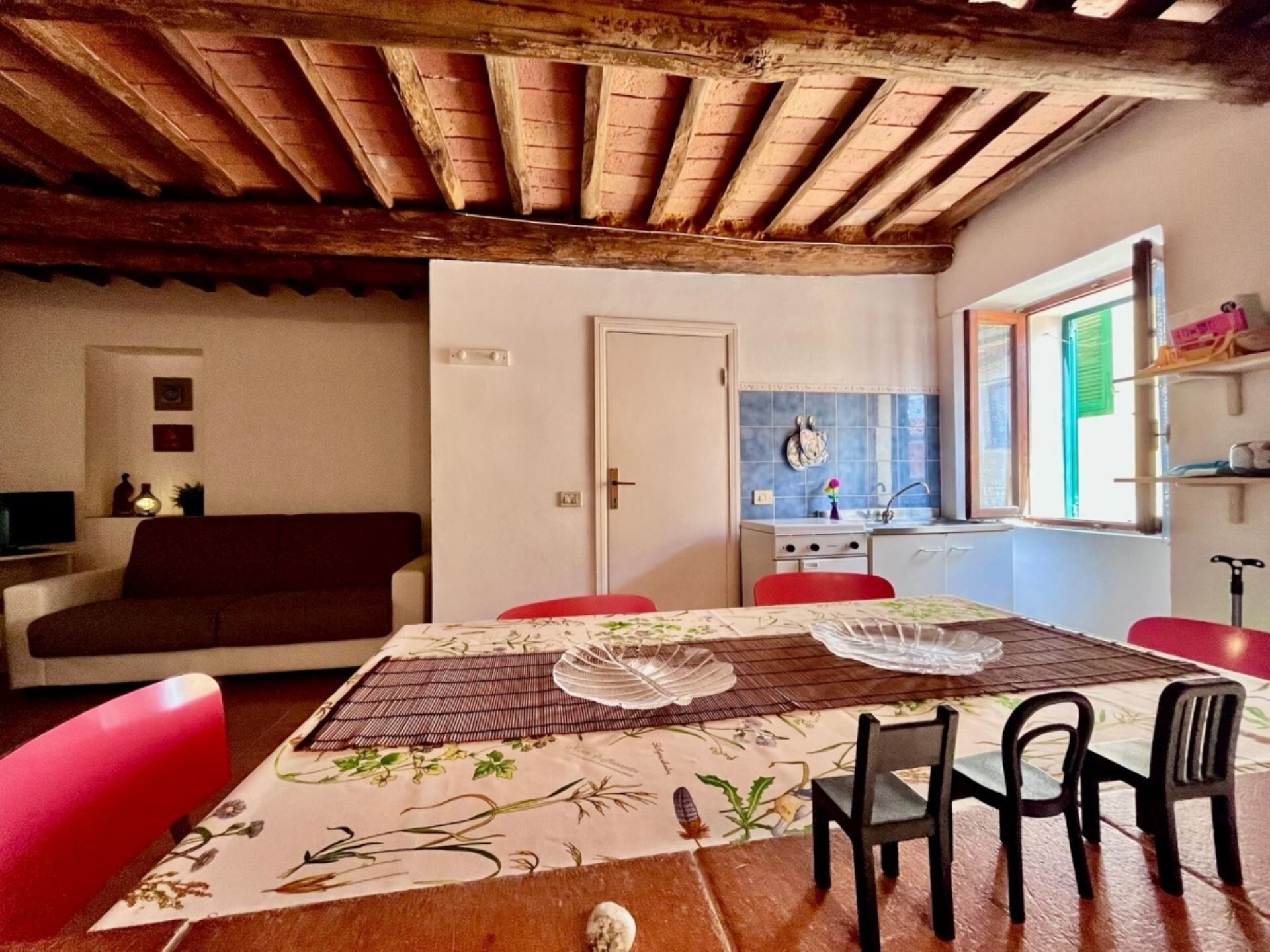 Un salon chaleureux avec un plafond en bois, une table à manger recouverte d’une nappe à motifs floraux et un coin cuisine avec une fenêtre.