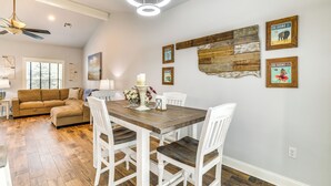 Un espace repas avec une table en bois et des chaises blanches, un ventilateur de plafond et des œuvres d’art accrochées au mur.