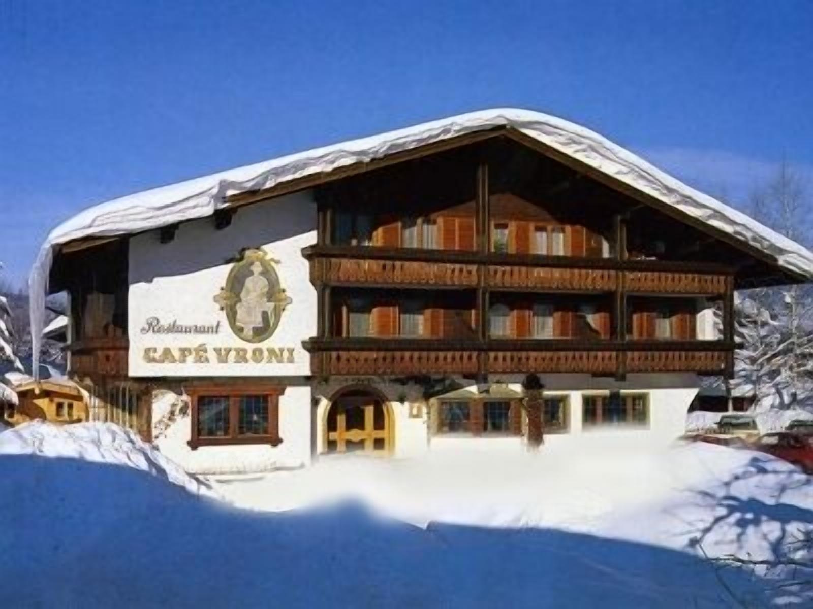 Un edificio in stile alpino tradizionale con un'insegna che recita "Restaurant Café Vroni". Presenta una facciata in legno, diversi balconi e un tetto coperto di neve.