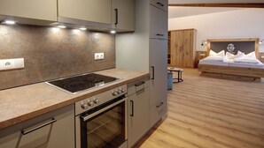 Una cocina moderna con estufa, horno y armarios integrados. Se observa una mesa de comedor de madera y, al fondo, una cama con cabecera de madera.