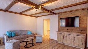Un salon moderne avec un plafond en bois, un canapé gris agrémenté de coussins bleus, une petite table ronde et un téléviseur fixé sur un mur en bois.