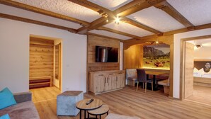 Une pièce chaleureuse et bien éclairée, avec des poutres en bois, un téléviseur à écran plat, un coin repas agrémenté d’une fresque représentant des montagnes, et un canapé avec des coussins.