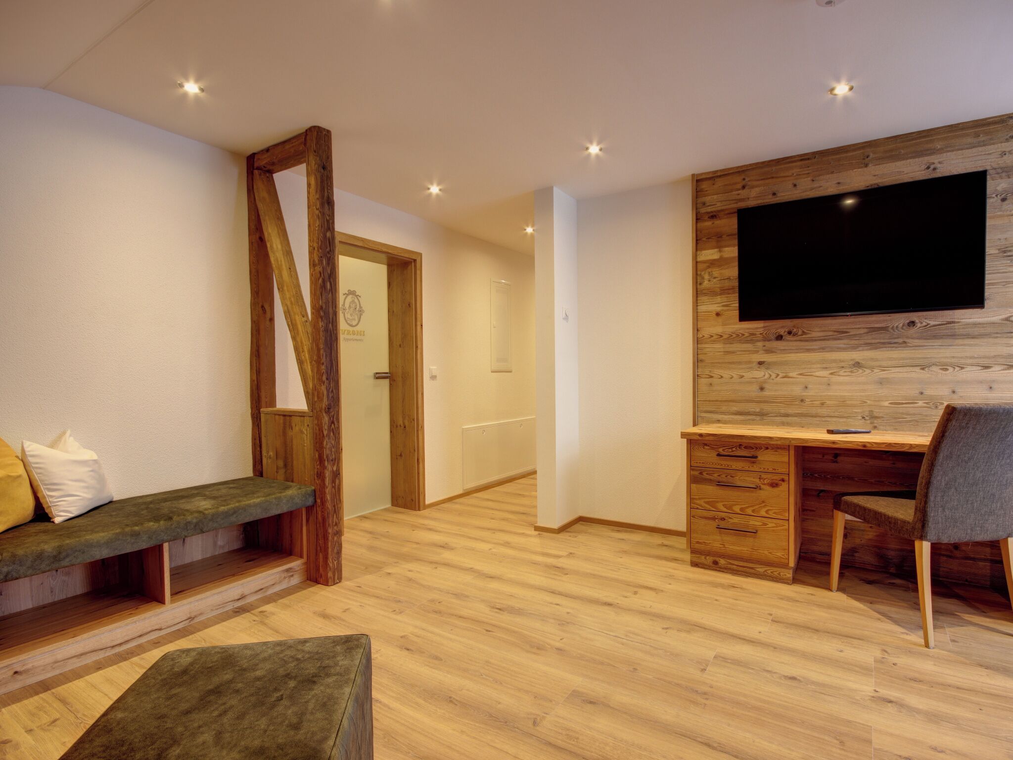 Une chambre moderne avec un bureau en bois, un téléviseur fixé à un mur en bois, un banc et une porte ornée d’un logo.