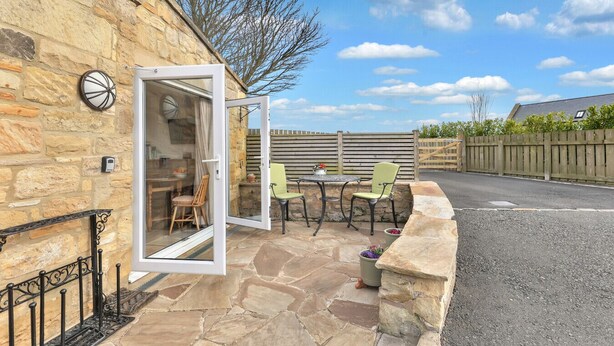 Un bâtiment en pierre doté d’un patio avec une table et des chaises, une porte donnant sur un espace intérieur, et une clôture en bois en arrière-plan.