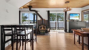 A modern living room with a wooden ceiling, a fireplace, a dining area with chairs, a television, and a view of the outdoors. - Havre du Huit (Adstock)