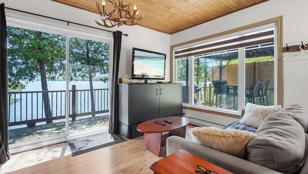 A room with a wooden ceiling, a television on a cabinet, a sofa with cushions, and a large window with a view of a beach and trees. - Havre du Huit (Adstock)