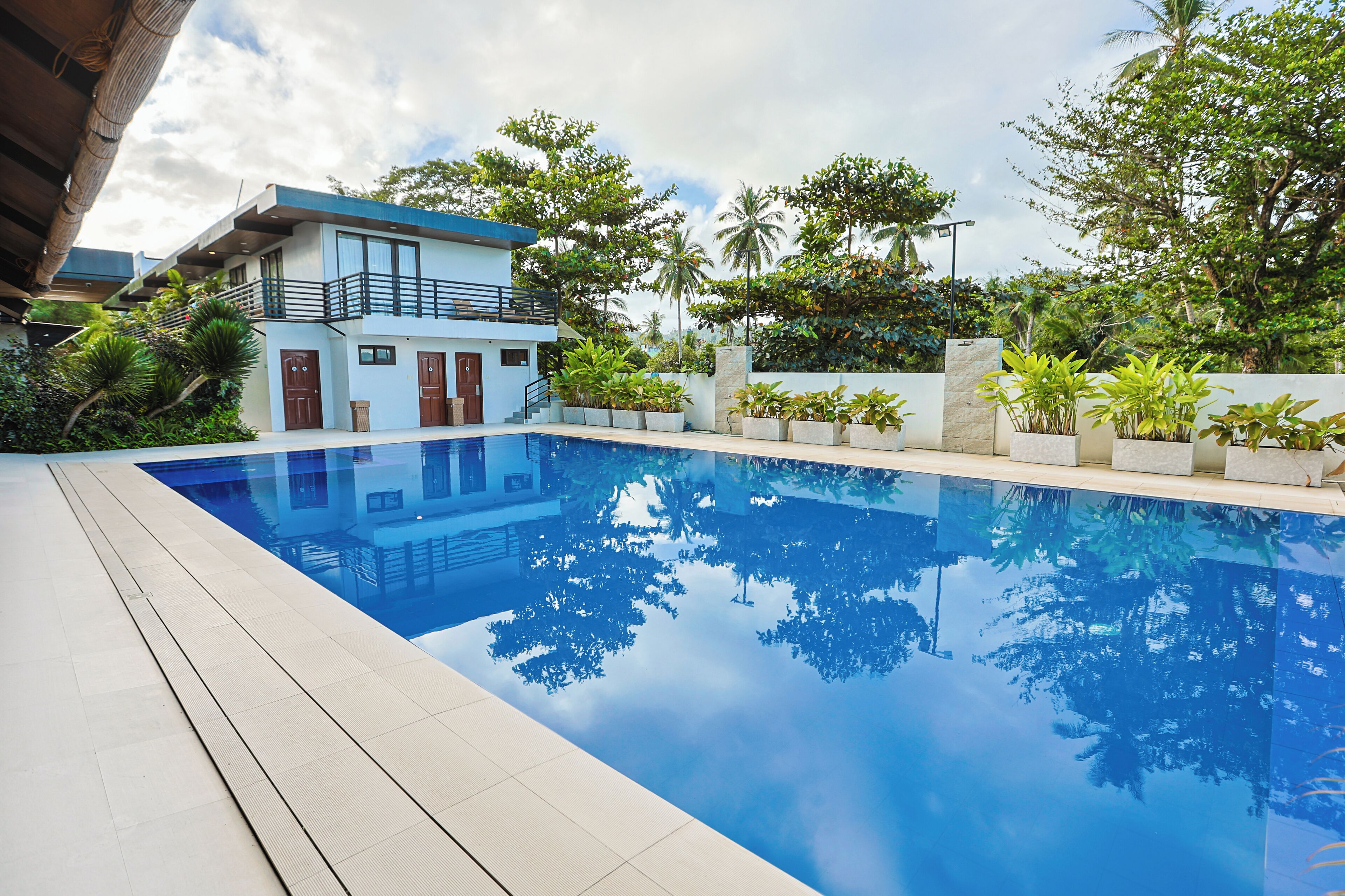 A modern pool area with a clear blue pool, surrounded by lush greenery and a two-story building with balconies.