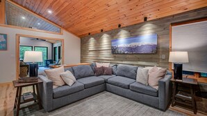 A cozy living room with a gray sectional sofa, wooden ceiling, and a mountain landscape painting. - Family-Friendly Chalet: Hot Tub, Games, Fire Pit (Jackson)