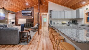 A modern interior with a kitchen, dining area, and living room, featuring wooden floors, a fireplace, and a high ceiling. - Family-Friendly Chalet: Hot Tub, Games, Fire Pit (Jackson)