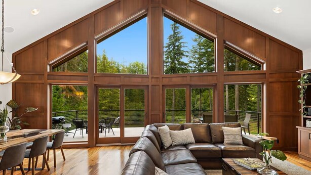 A spacious living room with a large sectional sofa, a dining area with a wooden table and chairs, and large windows offering views of a forested area.