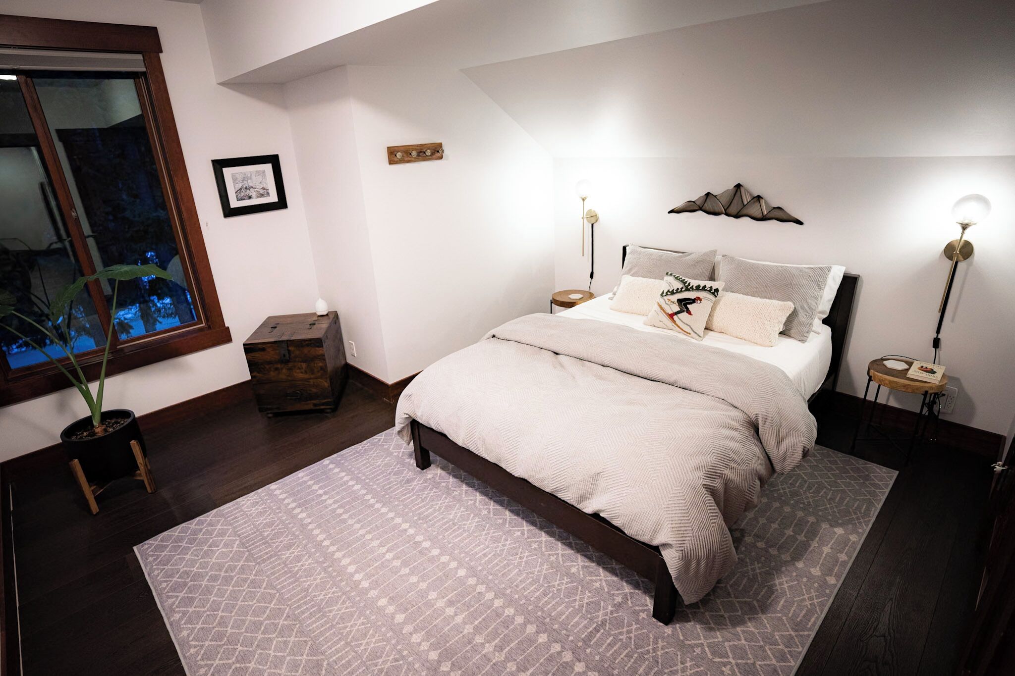 Ein Schlafzimmer mit Bett, Nachttischen, einem Topf mit Pflanze, einem gerahmten Bild und einem Fenster mit Ausblick.