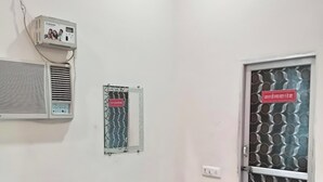 A small room with a bed, a desk, a wall-mounted air conditioner, a window with a metal grill, and a door with a sign. - Prem Bhawan Guset House (Danta Ramgarh)