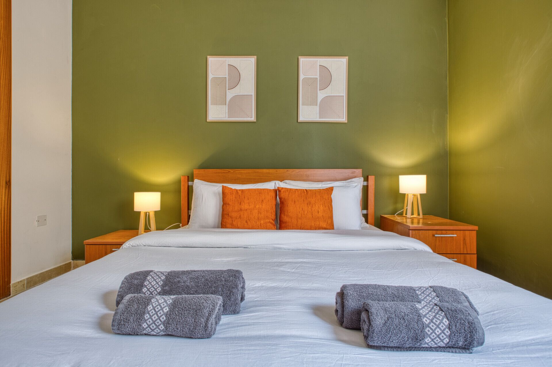 A neatly made bed with white and orange pillows, two bedside lamps, and two towels on the bed.