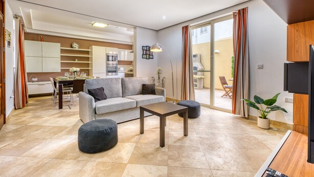 Ein modernes Wohnzimmer mit grauem Sofa, einem Couchtisch und einem schwarzen Hocker. Im Hintergrund befindet sich eine Küche mit weißen Schränken und ein Essbereich mit Tisch und Stühlen.