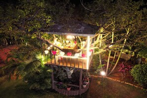 Una casa en el árbol con plataforma de madera, rodeada de vegetación exuberante y plantas en macetas.