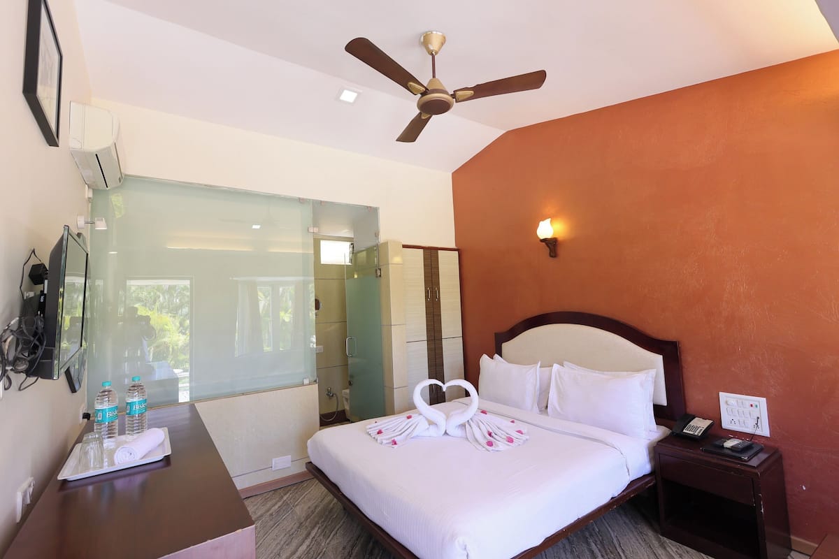 A hotel room with a bed, a bedside table with a phone and a lamp, a ceiling fan, and a glass shower enclosure.