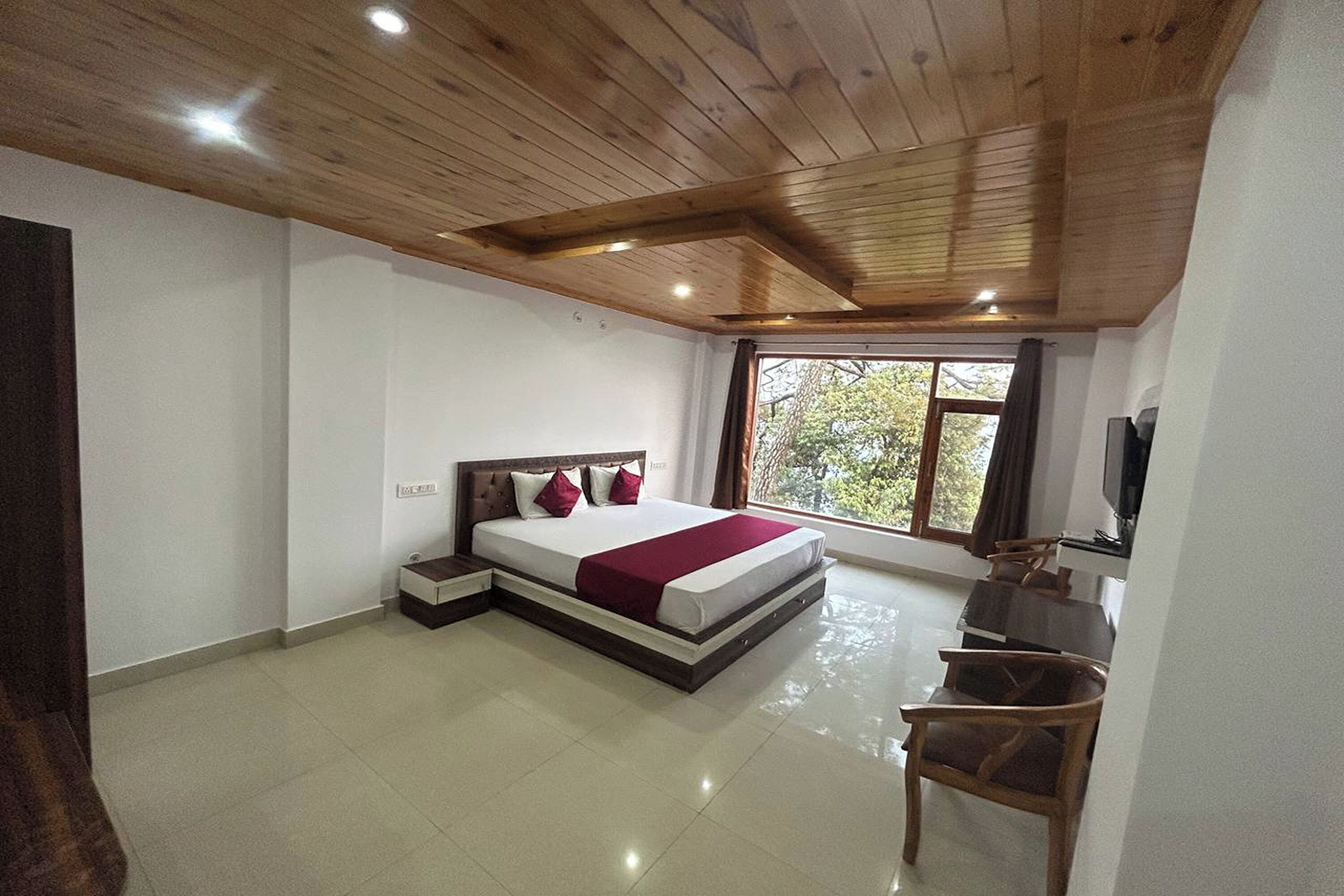 Un dormitorio con techo de madera, una cama con manta roja, un escritorio con silla y una ventana con vistas a árboles.