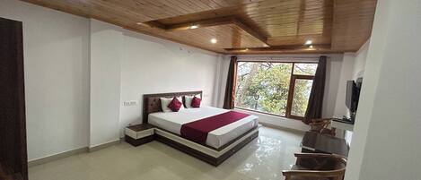 Un dormitorio con techo de madera, una cama con manta roja, un escritorio con silla y una ventana con vistas a árboles.